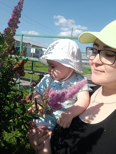 A woman wearing a yellow cap and black tank top, with glasses, holds a baby wearing a white sun hat and light blue t-shirt in her arms. The baby is looking at pink flowers on a plant. They are outside in a grassy area with buildings and a green fence in the background under a blue sky with white clouds. The scene appears to be in or near the city of Sergiyev Posad, Russia.