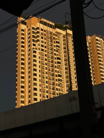 This media file captures a low-angle, upward view of a modern, cream-colored building with numerous windows, set against a dark, overcast sky. The building's facade features repeating horizontal bands of light brown and cream, creating a rhythmic pattern. The windows are mostly dark, with some reflecting the dim light. The building appears to be very tall, with multiple stories visible.
The perspective is unusual, as the building is viewed from below and is tilted. A thick, dark beam runs horizontally across the upper portion of the image, obscuring part of the building and casting a shadow. Below this beam, the building's reflection is visible, mirroring the structure above, suggesting the image might have been taken in relation to a reflective surface like water or glass, or it could be an optical illusion due to the angle.
Several dark, thin wires are strung diagonally across the frame, adding to the urban feel. A small red sign with white text is visible on the underside of the horizontal beam. The text on the sign is partially obscured but appears to read "BHT 3761".
The lighting suggests it is either early morning or late evening, with the sun low in the sky, casting a warm glow on the building. The dark sky and absence of bright sunlight imply potentially cloudy or stormy weather. The presence of power lines and the architectural style of the building are indicative of an urban environment, possibly in Bangkok, Thailand, as suggested by the location context. There are no people or clear activities depicted; the focus is on the architecture and the abstract interplay of light, shadow, and reflection.
