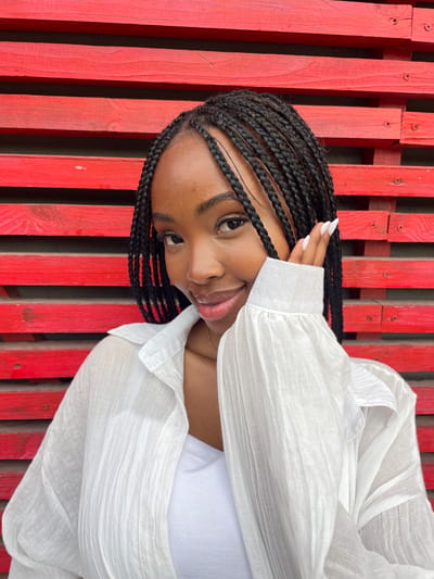 A young Black woman with braided hair and white acrylic nails is captured in a medium shot, leaning forward with her left hand near her cheek. She is wearing a white, open-collared shirt over a white tank top. The background is a vibrant red wall made of horizontal wooden planks. The image does not provide any information about the location being in Kazan, Russia.