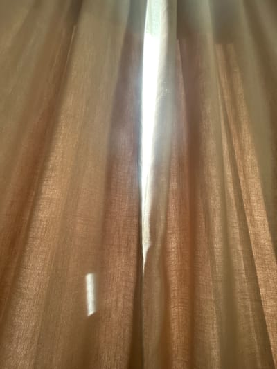 The image shows a close-up view of translucent, light brown curtains with vertical folds. Sunlight streams through a gap in the center of the curtains, creating a bright white vertical area. A rectangular reflection of light is visible on the left curtain. The scene depicts the interior of a room where light is filtering through window coverings. There are no people or specific landmarks visible to determine a location in Plovdiv, Bulgaria.