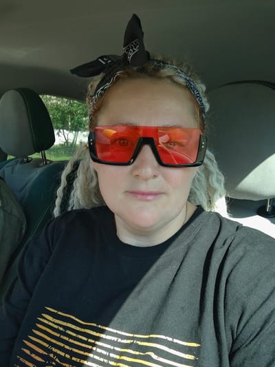 A woman with blonde curly hair and a black bandana is inside a car. She is wearing oversized red-tinted sunglasses and a black t-shirt with a yellow striped graphic. The interior of the car, including headrests and part of the dashboard, is visible. Sunlight streams in from the right, casting shadows on the woman and the car's interior. Outside the car, trees and greenery are visible through the windows. The location is the city of Марковский, Russia.