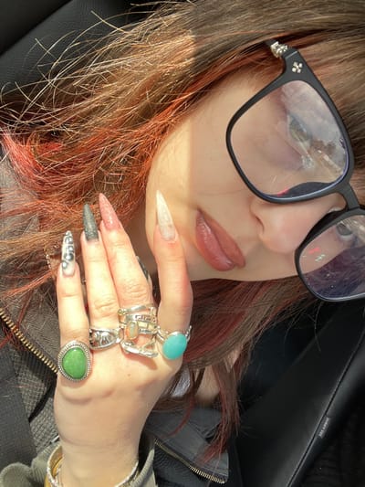 A close-up, high-angle portrait shows an individual inside a vehicle. The subject, likely female, has reddish-brown hair with lighter red highlights, strongly illuminated by direct sunlight. She wears black-framed glasses featuring silver cross embellishments on the temples; reflections in the lenses display blurred outdoor elements, including trees and what appears to be a red traffic light. Her lips are visible, coated in a brownish-pink substance.

The subject's left hand is prominent in the foreground, adorned with very long, pointed (stiletto) acrylic nails. The index finger nail is off-white, the middle finger nail is translucent pink with glitter, the ring finger nail is dark grey with glitter, and the pinky finger nail is white with black swirl patterns. Numerous rings are worn on this hand: a large oval green stone set in silver on the index finger; two silver rings on the middle finger, one featuring an oval iridescent orange stone and a geometric band, the other an abstract silver band; and a large oval turquoise stone set in silver on the ring finger. A gold-toned bracelet is partially visible on the wrist.

The environment is identified as the interior of a car, characterized by black upholstery and a visible "SRS AIRBAG" label on a panel to the subject's right. The scene is depicted during daylight hours. While the image is situated in the city of Raanana, Israel, specific geographic markers within the city are not discernible from the visual content.