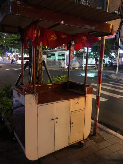 This media file shows a street scene at night in Da Nang, Vietnam. The primary focus is a food cart or stall with a white cabinet on the right side, featuring two drawers with silver knobs. A red metal frame supports a canopy or awning on the left, which is adorned with two Vietnamese flags, each featuring a yellow star on a red background. These flags appear to be made of fabric and are slightly tattered.
Behind the cart, a red and white striped barrier, likely a gate or bollard, is visible, extending horizontally across the scene. The ground is paved with rectangular stones on the right, and a wet asphalt road with a pedestrian crossing is visible on the left. Streetlights illuminate the scene, reflecting on the wet surfaces, suggesting recent rain or a humid environment. Lush green foliage is present in the background, adding a touch of nature to the urban setting.
Further back, buildings with illuminated signs and windows can be seen, indicating commercial activity. A few people are visible in the distance, blurred by the low light and motion, suggesting the street is active at night. The overall impression is a typical street view in a Southeast Asian city after dark, with elements of commerce, daily life, and potentially national pride represented by the flags. The time of day is clearly night, and the wet surfaces imply damp weather conditions. There is no discernible text that can be read clearly from the image.