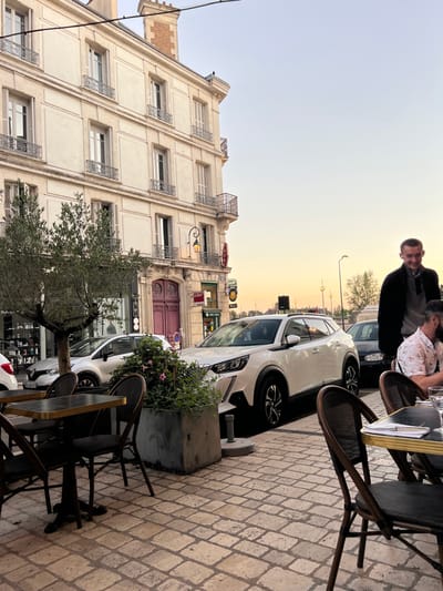This image depicts a street scene in Orléans, France, specifically on Rue des Capucins, identifiable by the presence of "La Riposte" establishment visible on the building. The scene captures a late afternoon or early evening ambiance, indicated by the sky's gradient from pale blue to warm orange at the horizon.
In the foreground, a cobblestone pavement extends across the lower portion of the frame. Several café tables with dark tops and dark chairs with woven seats are arranged on the left side, appearing unoccupied. A large, grey concrete planter containing green foliage and small pink flowers is positioned between the café seating and the street.
The midground features three parked vehicles. On the far left, partially obscured by an olive-like tree, is a light-colored Renault Clio hatchback. Dominating the center is a white Peugeot 2008 SUV, parked parallel to the curb, facing away from the viewer. Further down the street, behind the Peugeot, a dark-colored Volkswagen Golf is partially visible.
Two individuals are present on the right side of the frame. A man with a beard, wearing a patterned shirt, is seated at a café table, seen from the chest up, looking downwards to his right. Behind him, another man stands, dressed in a dark jacket over a grey garment, smiling slightly towards the camera.
The background is dominated by a multi-story, light-colored stucco building with classical European architectural elements, including multiple windows with ornate wrought-iron balconies and railings. The ground floor of this building features a prominent, ornate dark red double door. Below the first-floor balconies, a sign for "La Riposte" is visible. On the corner of the building, a red "ABA" sign is affixed. Streetlights are visible further down the street in the distance.