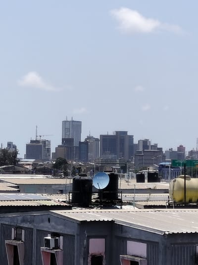 The image displays an elevated urban landscape in Lagos, Nigeria, under a hazy, light blue sky with scattered white clouds. In the immediate foreground, a concrete building with vertical corrugated metal siding features several rectangular openings, one containing an air conditioning unit. Its roof is composed of corrugated metal. On various foreground and mid-ground rooftops, multiple water storage tanks are visible: two prominent large black cylindrical tanks, one yellow cylindrical tank, and several smaller black tanks. A blue DStv satellite dish, bearing the DStv logo, is mounted on a stand next to one of the large black tanks. Several flat solar panels are present on other mid-ground rooftops. The distant background presents the Lagos Island central business district skyline, characterized by a cluster of approximately fifteen to twenty high-rise commercial and residential buildings with glass and concrete facades. A construction crane is visible near the left edge of the skyline, indicating ongoing development. No human figures or discernible activities are present within the frame.