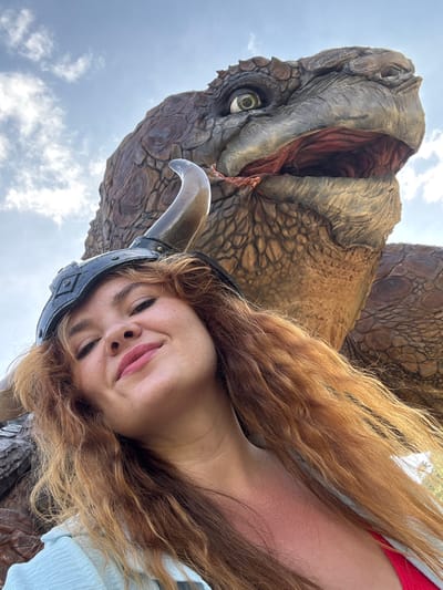 An outdoor, ground-level, close-up selfie shows a smiling young woman with long, wavy, red-blonde hair. She is wearing a black helmet with a horn and a light blue shirt over a red garment. Behind her, a large, reptilian creature, possibly a dinosaur or dragon, with scaly, brown skin and a wide-open mouth showing red interior and sharp teeth, is partially visible against a cloudy blue sky. The scene appears to be in an amusement park or a themed attraction.