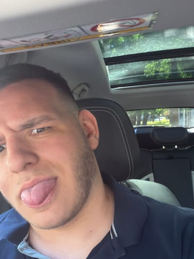 The image shows a close-up, selfie-style photograph taken from inside a vehicle. The subject is a young man with short hair, stubble, and his tongue sticking out. He is wearing a dark blue polo shirt. The car's interior is visible, including a sunroof with trees and foliage reflected in it, and black leather seats with white stitching. The sun visor in the car has a "no smoking" symbol visible. The image does not provide any specific visual cues to determine the location within the city of Plovdiv, Bulgaria.