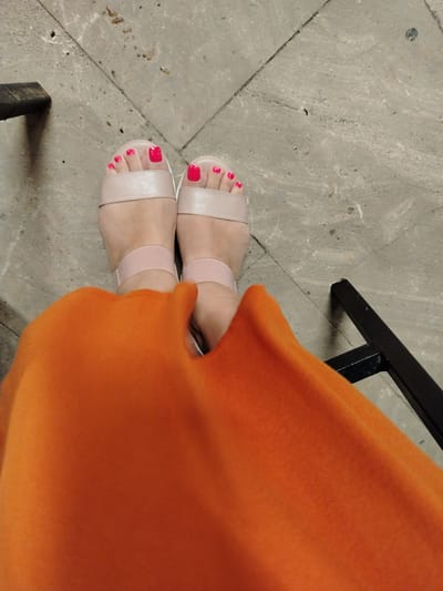 A pair of feet, adorned with bright pink nail polish and beige sandals, are positioned on a textured, light-colored stone surface. The feet are partially obscured by a vibrant orange garment, suggesting the person is wearing a dress or skirt. A black metal object, possibly part of a chair or table, is visible in the lower left and right portions of the frame. The lighting suggests an outdoor or well-lit indoor environment. The scene does not provide any specific visual cues to determine its location within Sumqayıt, Azerbaijan.