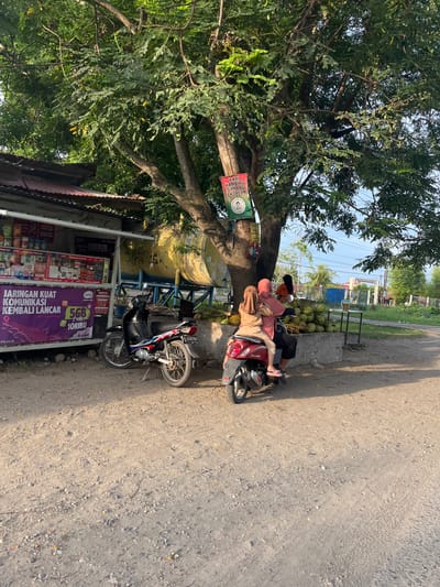 The image depicts a street scene in Lhokseumawe, Indonesia. Two individuals, a man and a woman wearing hijabs, are seated on a red scooter under the shade of a large tree. The woman is leaning back against the man, who appears to be reaching for something. The scooter is parked on a dirt road alongside a small roadside stall.
The stall has a corrugated metal roof and displays various items, possibly snacks or prepaid phone cards, behind glass. A prominent purple banner with text in Indonesian is attached to the front of the stall, advertising "JARINGAN KUAT KOMUNIKASI KEMBALI LANCAR" (Strong Network Communication Back to Smooth) and an offer from AXIS for 5GB of data for 10,000 Indonesian Rupiah for 2 days. A green sign with text and an image is also visible on the tree, reading "KARI KAMBING SIMPANG EK TREUN" (Curry Goat Crossroads Ek Treun), suggesting a local eatery.
In the background, a pile of coconuts is visible next to a concrete ledge, and further back, buildings and trees indicate a semi-rural or suburban environment. The sunlight suggests it is daytime, likely late afternoon given the long shadow cast on the road. The overall atmosphere appears relaxed, with the individuals taking a break from their journey or activity. The license plate of the scooter is partially visible, showing "BL 4483 ND".