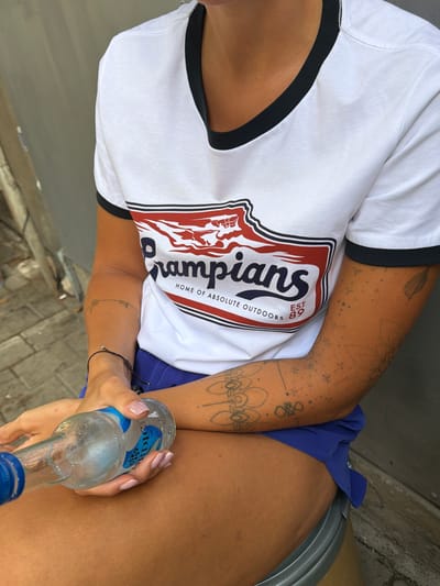 The image shows a close-up of a person holding a clear glass bottle of what appears to be a beverage. The person's arm, adorned with tattoos, is visible, and they are wearing a white t-shirt with a black trim around the collar and sleeves. The t-shirt features a graphic logo with the text " CHAMPIONS HOME OF ABSOLUTE OUTDOORS EST 89". The person is also wearing blue shorts. Their hand, with manicured nails, is gripping the bottle. The background suggests an outdoor setting, with hints of paving stones and a metallic structure. The lighting indicates it is daytime. The setting is identified as Tel-Aviv, Israel. The overall impression is casual and relaxed.