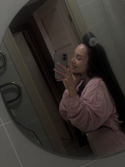 The image is a mirrored selfie taken in what appears to be a bathroom. A person with long dark hair, wearing a pink tracksuit, is holding a cell phone to capture the image. A hair roller is visible in the person's hair. The reflection reveals bathroom fixtures like a sink, a cabinet, and tiled walls. The photo has a muted, greyish tone. There is no indication of Kudrovo, Russia, within the image.