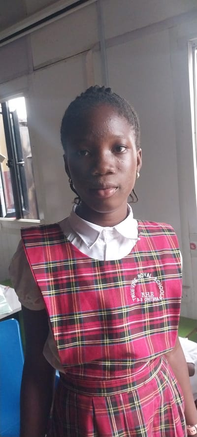 A young Black girl, likely of school age, is depicted from the chest up in an indoor setting. She is wearing a white collared shirt beneath a red, plaid jumper dress. The dress features a circular embroidered emblem on the chest that reads "TRUEVINE ROYAL SCHOOLS" with "RHE 3-3-3 ONITSHA" below it. Her hair is styled in braids, and she has small earrings in her ears. The background suggests a classroom or similar educational environment, with a window visible on the left side of the frame and a hint of blue furniture on the far left. The lighting is somewhat harsh, casting shadows and highlighting some shine on her skin. The overall impression is a posed, head-and-shoulders portrait taken within the Truevine Royal Schools in Onitsha, Nigeria.