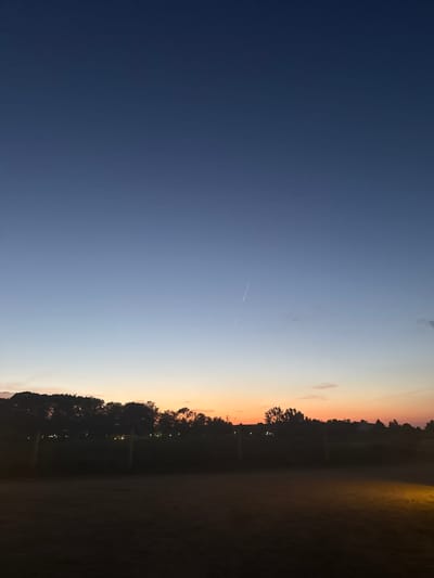The image depicts a twilight sky over the coastal village of Shkorpilovtsi, Bulgaria. The sky transitions from a deep blue at the top to lighter shades of blue and then to a warm orange and pink hue near the horizon, indicating sunset or sunrise. A faint contrail from an aircraft is visible in the upper half of the sky. Silhouetted against the colorful sky is a line of trees and vegetation, forming the distant treeline. Below the treeline, there are faint lights that appear to be from buildings or streetlights. In the foreground, the ground is dark and appears to be a grassy or dirt area, with a single spot of yellow light illuminating a small portion of it from the right side of the frame. The overall impression is one of a serene, natural landscape at the transitional time of day.