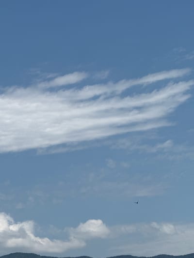 This is a photograph taken from a high vantage point, likely on a clear day with some scattered clouds. The upper two-thirds of the image are dominated by a clear blue sky, with a few wispy white clouds stretching horizontally across the frame. In the lower third, there are larger, more substantial white clouds and the dark silhouette of a mountain range in the distance, suggesting a natural landscape. A small, dark object, possibly a bird or an aircraft, is visible in the mid-right portion of the sky. The image does not provide any specific landmarks or features that would definitively place it in Tbilisi, Georgia.
