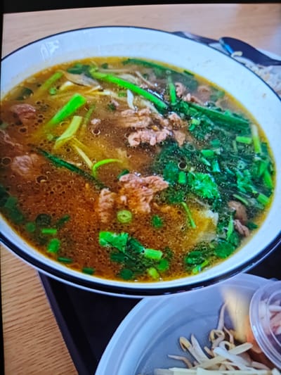 A close-up shot shows a steaming bowl of pho, a Vietnamese noodle soup, as the main subject. The soup is a rich brown color and is filled with thinly sliced beef, noodles, and an assortment of fresh green vegetables, including scallions and cilantro. The bowl itself is white with a black rim, placed on a dark tray that rests on a light brown, wooden surface. To the right of the soup bowl, a smaller, light blue plastic container is partially visible, holding bean sprouts and possibly some thinly sliced onions. The scene appears to be an indoor setting, likely a restaurant, given the presentation of the food. The lighting suggests it could be daytime, though the ambiance is warm and inviting, emphasizing the comforting nature of the dish. There are no people visible in the frame, and no visible text. The overall impression is one of a delicious and hearty meal, ready to be enjoyed.