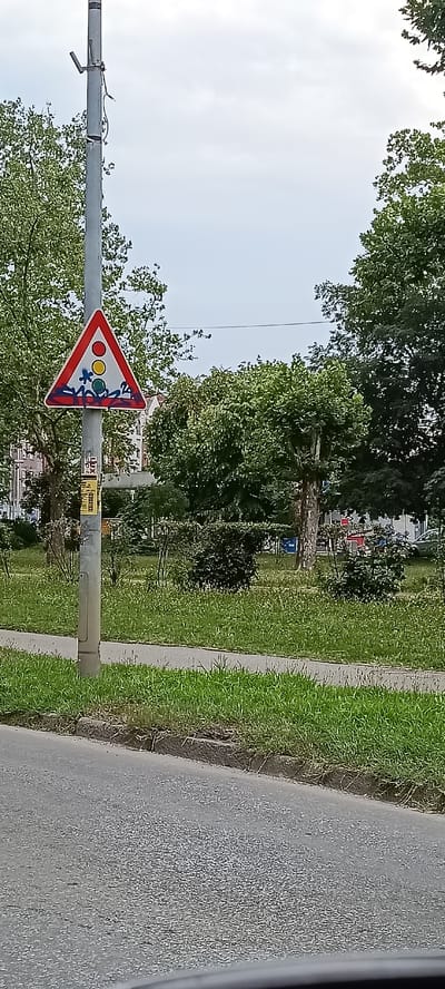 The image shows a traffic sign depicting a traffic light with red, yellow, and green circles. The sign also features a depiction of children playing, indicated by stylized figures. The sign is attached to a metal pole. In the background, there are trees, buildings, and a cloudy sky. A paved road is visible in the foreground. The scene appears to be in a park-like setting with grass and bushes. The traffic sign is graffitied with blue and red spray paint. This traffic sign is located in the city of Novi Sad, Serbia, specifically near the beginning of Bulevar Oslobođenja, close to the intersection with Novosadski Put.