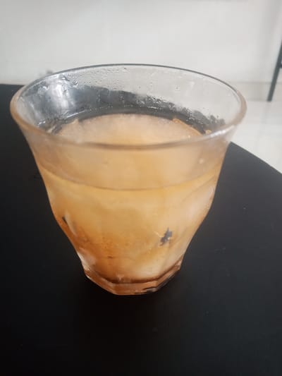A glass of iced tea sits on a black surface. The glass is partially filled with a light brown liquid and ice. Condensation is visible on the outside of the glass. The background is blurred and appears to be an indoor setting with a white wall and a dark chair leg visible. The image appears to be taken from a slightly elevated angle. The exact location in Depok, Indonesia, cannot be determined from the image alone.
