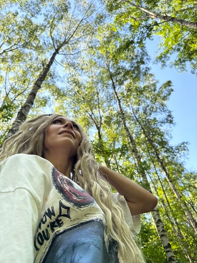 An outdoor, low-angle, medium shot captures a Caucasian woman with long, blonde, braided hair. She is wearing a white t-shirt with a graphic design. Her face is turned upwards and to the left, with her eyes looking in that direction and her mouth slightly open. Her right arm is bent, with her hand resting near her head, partially obscuring her hair. The background consists of tall birch trees with green foliage, against a clear blue sky, suggesting a forest or park setting in Rusanovo, Russia. The lighting is natural and bright, indicating daytime.