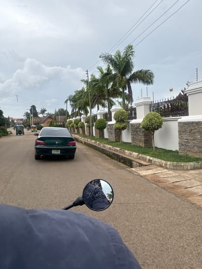 This media file captures a scene from a moving vehicle, likely a motorcycle given the perspective and the presence of a side mirror reflecting the rider. The image is oriented vertically, showing a street scene in Gura Top, Nigeria. 

The main subjects include a dark green sedan driving ahead on the paved road, and several smaller vehicles, including what appear to be auto-rickshaws or tricycles, further down the street. The roadside is lined with manicured green lawns and trees, including palm trees and intricately shaped bushes, suggesting a well-maintained residential or commercial area. A modern building with white walls, stone accents, and a decorative balcony is visible on the left side of the road. Power lines run overhead, typical of many urban and suburban areas.

The weather appears to be overcast with a bright sky, possibly indicating a cloudy day or the aftermath of rain, as evidenced by the reflection of foliage in the motorcycle's side mirror, which suggests it is wet. The time of day is likely daytime, given the ambient light. The scene conveys a sense of everyday movement and urban life in Nigeria.

Visible text includes a license plate on the green sedan, which reads "SG 206 DU". There are also markings on the license plate that are difficult to discern clearly.