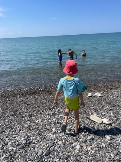 In Giachrypsh, Abkhazia, a sunny day unfolds at a pebble beach. The turquoise sea, calm and clear, stretches to the horizon under a bright blue sky. In the shallow water, four individuals are present. A woman in a blue swimsuit stands on the left, facing right. Next to her, a shirtless boy in dark swim trunks faces left, his arm extended as if interacting with the woman. Further back, a woman in a black swimsuit is submerged up to her chest. Another individual, barely visible, is in the water behind the boy.
On the shore in the foreground, a child wearing a red bucket hat and a blue and yellow swimsuit faces away from the camera, walking towards the sea. The child holds a small white object in their right hand. To the child's right, two pairs of white flip-flops lie on the pebble-strewn beach. The pebbles are a mix of gray, brown, and white, smoothed by the water. The overall scene depicts a relaxed summer day at the seaside.