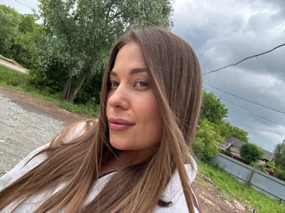 A young woman with long, light brown hair takes a selfie outdoors in Chernushka, Russia. She is wearing a white hoodie. The background includes trees, a dirt road, and a grey, overcast sky. In the distance, there are houses and a metal fence.