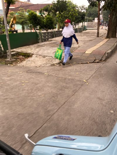 The image shows a person walking on a sidewalk in the city of Bandung, Indonesia. The person is wearing a white hijab and a blue shirt. The person is carrying a green bag and is walking towards the right. The image was taken from a moving vehicle, likely a motorcycle, which is visible in the bottom of the frame. The sidewalk is cracked and uneven. There are trees and bushes lining the sidewalk. The location is in the city of Bandung, Indonesia, based on the coordinates provided. The coordinates suggest that the image was taken in a residential or commercial area.
