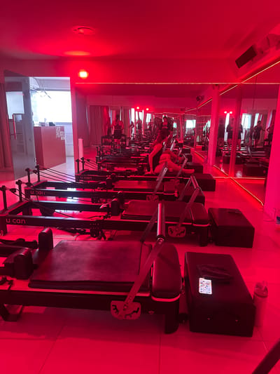 The image shows the interior of a brightly lit studio. The room is filled with Pilates reformer machines, arranged in rows. Each reformer has a sliding carriage, springs, and foot bar. In the background, a large mirror reflects the rows of reformers and the red lighting. The red lighting illuminates the room, casting a strong hue over everything. Some of the machines are in use by people who are wearing black clothing. The window in the background shows some signs for 'HOT PILATES', and 'U CAN'. The signage indicates that the location is most likely in the city of Puebla, Mexico.