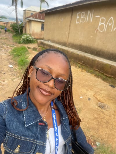 In the city of Rafin Par, Nigeria, a young woman with braided hair and sunglasses smiles for a selfie. She is wearing a denim jacket over a white top and a blue lanyard that reads "University of Jos (Stu." The background shows a dirt road and buildings, with some greenery and a palm tree. Graffiti is visible on one of the buildings. The overall scene is outdoors, in what appears to be a residential or semi-urban area.