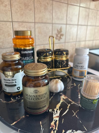 The image shows a close-up of a dark marble tray with several items on it, placed on a surface with beige square tiles. The items include three brown glass bottles of supplements, one large orange bottle of supplements, a white bottle of supplements, a whole garlic bulb, and a clear plastic container filled with toothpicks. The brown bottles are labeled "Gentle Zinc + Vitamin A", "Magnesium Glycinate", and another is mostly obscured by the others. The large orange bottle has "ShiffaHome OMEGA" printed on it. The white bottle is labeled "D3 + K2". The toothpick container has Russian text and the number "190" visible. In the background, two decorative black and gold containers are visible on a gold rack. The scene appears to be a domestic setting, possibly a kitchen counter. There are no people visible in the image. The lighting suggests an indoor environment. The overall impression is one of health and wellness products.