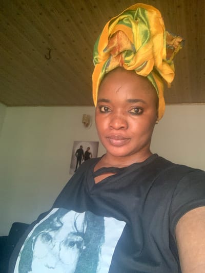 An indoor portrait depicts a Black female individual, presumably located in Barakin Akawo, Nigeria. The subject faces forward, looking directly into the camera with a neutral expression. She wears a multi-colored fabric headwrap, predominantly yellow and green, styled voluminously on her head. A light-colored scar is visible on her forehead, above her left eye. Her eyes are accentuated with dark eyeliner, and a gold stud earring is visible in her right ear. She is dressed in a black V-neck t-shirt with a white graphic print on the front depicting a face with round glasses. The background features a light-colored wall with a wall sconce and a framed picture showing two silhouetted figures. Part of a dark furniture item is visible in the lower left. The ceiling is constructed from light brown wooden planks, with a dark, J-shaped hook present in the upper left quadrant.