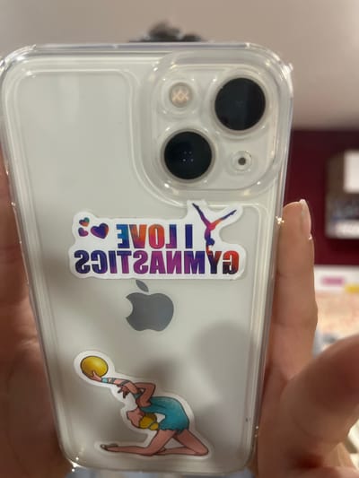 A person's hand holds a clear iPhone case with stickers on the back. The stickers include colorful hearts, the text "I LOVE GYMNASTICS" in a gradient of blue, purple, and red, a silhouette of a gymnast in a pose, and a sticker of a gymnast in a blue leotard holding a yellow ball. The Apple logo is visible below the "I LOVE GYMNASTICS" sticker. The background appears to be an indoor setting, possibly a room with soft lighting and some blurred objects. There is no information about the city of Moreni, Romania, in the image.