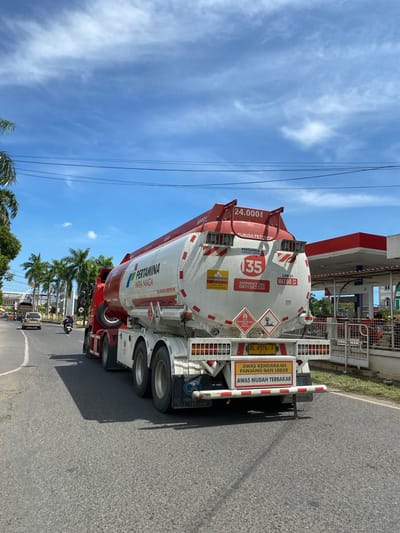 The image captures a Pertamina fuel tanker truck driving on a paved road in Lhokseumawe, Indonesia. The truck, red and white, has "PERTAMINA PATRA NIAGA" branding and a capacity of "24.000 L". It displays hazard symbols for flammable liquids and has a license plate reading "BL 9576 F". Text on the back warns "AWAS KENDARAAN PANJANG DAN LEBAR" (Beware of long and wide vehicles) and "AWAS MUDAH TERBAKAR" (Beware of flammability). To the right of the truck is a Pertamina gas station with red and white canopy, pumps, and a fence. In the background, palm trees line the street, and other vehicles, including a silver car and a motorcycle, are visible on the road. The sky is blue with scattered white clouds, and power lines are strung across the upper portion of the image. The scene appears to be daytime, with bright sunlight casting shadows.