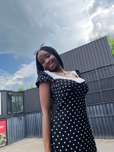 A young Black woman with braided hair, wearing a black and white polka dot dress with lace trim and sunglasses on her head, is pictured outdoors. She is smiling at the camera. Behind her are large, dark shipping containers stacked up, with some trees visible in the distance. The sky is overcast with scattered clouds. This scene is not located in the city of Kazan, Russia.