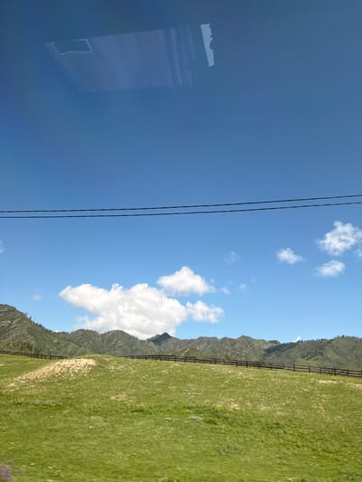 The image shows a pastoral landscape under a bright blue sky with scattered white clouds. In the foreground, a vibrant green, grassy hill slopes upwards, with a wooden fence running horizontally across its middle. Beyond the hill, a range of green, tree-covered mountains forms the horizon. Two dark electrical wires are stretched across the upper portion of the sky. In the top section of the image, a reflection is visible on what appears to be a window, showing a white, rectangular shape and vertical lines. The scene is indicative of the natural beauty found in rural areas like Tuekta, Russia.