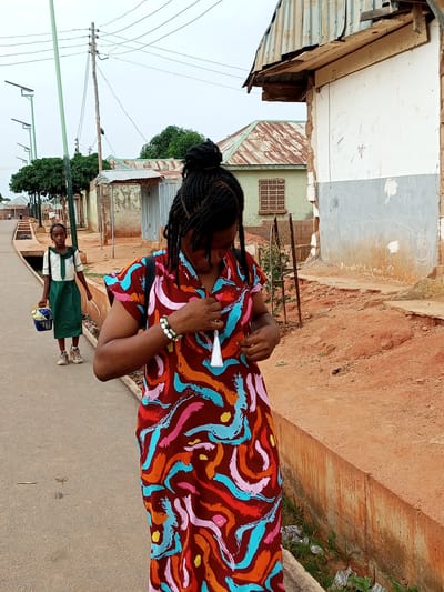 In Tempure, Nigeria, a young girl in a school uniform walks on the left side of a paved road, carrying a basket and a white object. A woman in a colorful dress stands on the right, adjusting her attire. The scene unfolds in a dusty, urban environment characterized by unfinished buildings with corrugated metal roofs, utility poles with overhead wires, and sparse vegetation. The road is flanked by a concrete drain on the right, filled with debris. The sky is overcast, suggesting a cloudy day.