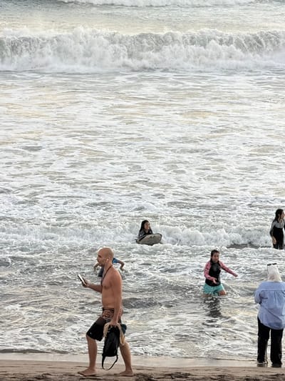 In North Kuta, Indonesia, a beach scene unfolds with several individuals in the shallow ocean and on the shoreline. In the foreground, a bald man in patterned swim trunks walks on the sand, holding a smartphone in his left hand and a dark bag with rope in his right. He is wearing earbuds. Further out in the water, a young girl in a pink and black rash guard and turquoise shorts kneels, looking towards the left. To her right, a woman wearing a blue striped shirt and black pants stands on the sand, facing the ocean. In the mid-ground, a young girl in a dark patterned rash guard sits on a surfboard, while another young girl in a dark full-body swimsuit stands to her right, also in the water. The ocean is choppy with significant wave activity and foam. In the background, large waves crash. The overall lighting suggests it is either early morning or late afternoon.