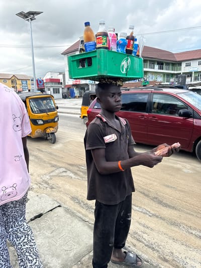 In Epe, Nigeria, a young boy balances a green crate filled with various beverage bottles on his head. He is dressed in a dark polo shirt and dark trousers. In his hands, he holds several folded banknotes, appearing to be engaged in a transaction. To his left, another person, partially visible, wears a pink t-shirt with a cartoon bear print and patterned trousers. Behind the boy, a red van is parked on a sandy road. Further in the background, a yellow auto-rickshaw is visible, along with buildings lining the street under an overcast sky. The overall scene depicts a typical street vendor environment in the city.