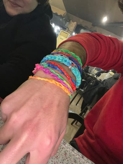 A person's arm adorned with multiple colorful, braided rubber band bracelets. The bracelets vary in color, including green, light blue, red, blue, pink, and orange. One bracelet features a single clear bead. The person wears a red long-sleeved shirt. The background includes a dark figure wearing a black shirt, a chair, and a patterned surface. The scene appears to be indoors with bright lighting.
