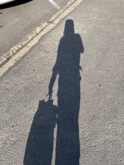 A shadow of a person is cast on a gray asphalt surface in Palaiseau, France. The shadow suggests the person is wearing a hat, a shirt, and long pants. They are holding an object, possibly a bag or briefcase, in their right hand. The asphalt surface is marked with a painted yellow line near the left edge of the frame. A small portion of a black tire is visible in the bottom left corner of the frame, which may indicate the presence of a vehicle. The light source is positioned at an angle to the person, casting a long shadow. The shadow indicates it is either early morning or late afternoon.