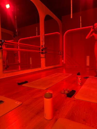 The image shows a room interior, possibly a fitness studio, illuminated by strong red light. In the reflection of a mirror, a person can be seen lying on a surface. Two exercise mats are present, along with various objects including weights, a water bottle labeled "owala," and a phone. A structure with wooden poles and metal bars is also visible. The number "11" is seen on a mat. The specific location is not evident from the image.