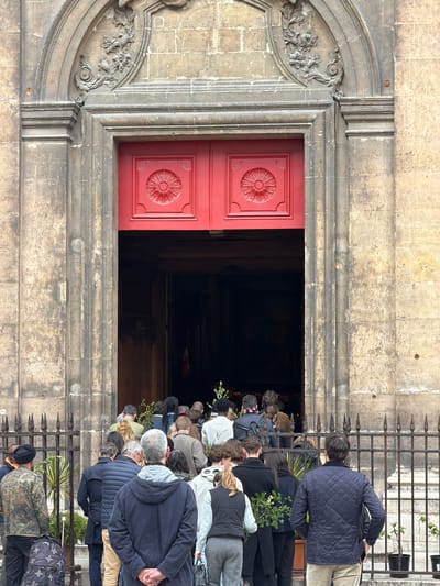 A street-level, medium shot captures a narrow, vertical view of an ornate building in Paris, France. A crowd of people in contemporary clothing stands on stone steps leading to a pair of partly open red doors. The doors are decorated with flower designs and set into a weathered stone frame with carved floral details. The stonework shows signs of age and discoloration. A black, spiked fence is set to the right of the frame. The people appear to be queuing. The lighting suggests a daylight setting.