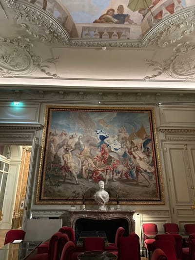 This image captures a lavish interior of a grand historic building, specifically identified as likely the Hôtel de Crillon located on Place de la Concorde in the 8th arrondissement of Paris, France.
The room features a high ceiling adorned with intricate white rococo stucco moldings and a central painted fresco depicting classical figures on a trompe l'oeil balustrade, observed from below. A small, white spherical security camera is mounted on the ceiling.
The primary visual focus is a monumental, richly woven tapestry, approximately 4-5 meters wide, framed in gold, which covers a significant portion of the main wall. This tapestry illustrates a chaotic historical or mythological battle scene, featuring numerous figures on horseback and foot engaged in combat, wielding weapons and flags, set against a dynamic sky. Prominent colors include reds, blues, whites, and muted earth tones.
Centered beneath the tapestry is an ornate marble fireplace with a reddish-brown and white veined mantlepiece. Perched on the mantle is a white marble bust of a woman, flanked by two dark, stylized decorative objects.
The foreground contains a seating area furnished with multiple armchairs upholstered in deep red velvet. These chairs are arranged around small tables with dark, reflective surfaces, suggesting a lounge or formal waiting area.
To the left, a partially open doorway leads to an adjoining hallway, revealing marble-clad walls and a green illuminated exit sign. The right side of the room features paneled walls with classical pilasters, consistent with the overall opulent architectural style.