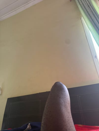 A first-person perspective shot of a person lying in bed in Narayi, Nigeria. The image shows a headboard of a bed, a wall, and a portion of the person's leg. The wall is a pale yellow color and appears to be textured. There are curtains in the background. The person is wearing a blue pajama bottom. The scene is lit by natural light coming from a window.