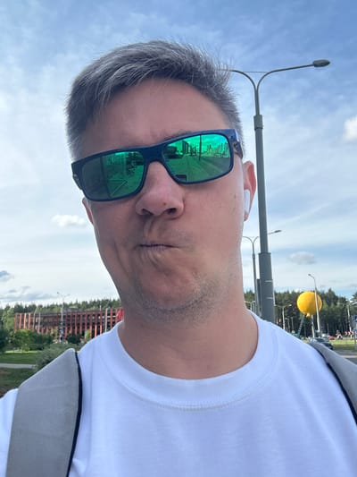 An adult male with short, grey hair and light stubble poses for a selfie outdoors on a partially cloudy day in Бараўлянскі сельскі Савет, Belarus. He is wearing a white crew-neck shirt and a grey backpack, visible on his left shoulder. His lips are pursed in a pouting expression, and his eyes are obscured by dark-framed sunglasses with reflective green lenses. A white wireless earbud is visible in his right ear. The sunglasses reflect a street scene featuring buildings, a road with white markings, and a bicycle. In the background to the left, a multi-story building with a reddish-brown slatted facade is visible behind green trees. To the right, tall grey streetlights with double arms stand against the blue sky, alongside more green foliage. A prominent, large yellow spherical object, possibly a decorative balloon or art installation, is visible on a tall pole in the distance, with what appears to be a ladder leaning against its support structure.