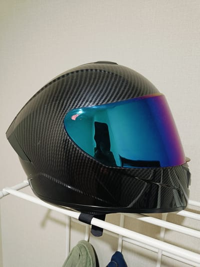 A black carbon fiber motorcycle helmet with a blue-to-purple iridescent visor is placed on a white metal rack against a textured off-white wall. The helmet is positioned on its side, with the visor facing forward and slightly upwards. The visor reflects a dimly lit interior, showing a silhouette of a person. The rack has multiple horizontal bars and a strap wrapped around one of the bars. Two pieces of clothing, one green and one dark blue, are hanging from the lower bars of the rack. The overall scene suggests an indoor setting, possibly a room or a storage area. There is no visible indication of the location being in Krasnodar, Russia.