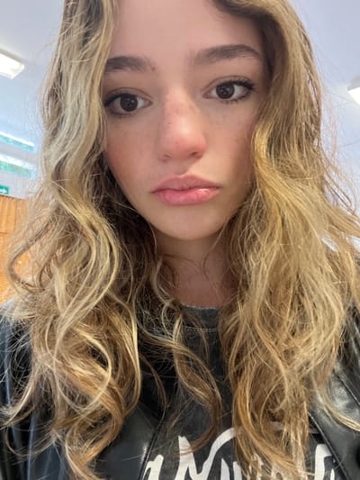 A close-up, angled portrait features a young person, likely taken as a selfie indoors in Puebla, Mexico. The person's head is significantly tilted or the image is rotated, showing them looking towards the viewer from the side. They have long, wavy, light blonde and golden-brown hair that frames their face and extends across much of the frame. Some darker brown strands or roots are visible.
The person's face is prominent, featuring brown eyes with long lashes, a nose with some visible freckles, and full, light pink lips. Their complexion appears clear, with a few light freckles on their cheeks and nose. Their eyebrows are natural and well-defined.
They are wearing a black, possibly leather or faux leather, jacket. A white design or pattern is partially visible on the right side of the jacket, behind the hair. The background is blurry and light-colored, suggesting an indoor setting with ample ambient light, possibly from a window or overhead lighting. No specific time of day is indicated beyond general daylight conditions. There is no visible text in the image. The overall mood is casual and direct.