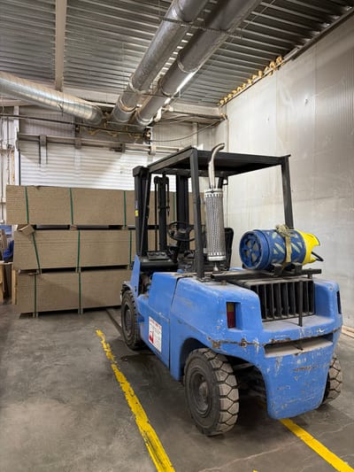 A blue forklift is parked on a concrete floor marked with yellow lines in a warehouse. The forklift has a black overhead guard and a metal exhaust pipe. A blue and yellow object is attached to the rear of the forklift. To the left of the forklift, stacks of flat, rectangular objects are piled high. The warehouse has a corrugated metal ceiling and walls. Ducts are visible on the ceiling. The scene is likely located in an industrial area of Sliven, Bulgaria. There are no people visible in the image.