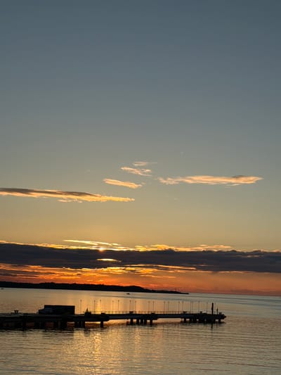 A sunset over the Baltic Sea near Tallinn, Estonia, casts a warm glow on the sky and water. The sun, partially obscured by dark clouds, is visible as a bright orb near the horizon. A pier extends into the water, with lampposts lining its edge. A truck and shipping container are present on the pier, suggesting a port or industrial area. The water reflects the vibrant colors of the sunset, creating a shimmering effect. The sky is a gradient of blue and orange, with wispy clouds scattered throughout. The scene evokes a sense of tranquility and natural beauty.