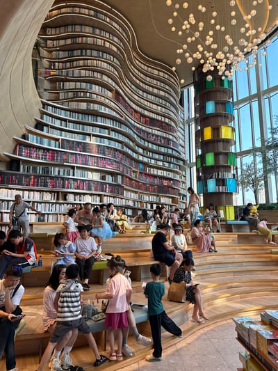 The image depicts the interior of the Zhongshuge Bookstore in Liangjiang, China. The bookstore features floor-to-ceiling bookshelves arranged in a wave-like pattern. Many people, including adults and children, are seated on the stepped, wooden floor in front of the bookshelves. Some are reading, while others are looking at their phones or interacting with each other. Above the bookshelves, there are cylindrical structures with colored sections (yellow, green, blue). A cluster of spherical lights hangs from the ceiling near the top left of the image. The architecture includes a glass roof, providing natural light to the interior.