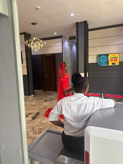 In a modern interior setting, possibly a public building or event venue in Abuja, Nigeria, a young woman adorned in a striking red traditional outfit, including a veil, stands facing left, her gaze directed towards something off-camera. She is positioned near a dark wooden door. In the foreground, a security personnel, facing away from the camera, sits behind a metal detector with a grey surface. They are wearing a white short-sleeved shirt and dark trousers. The background features a television screen displaying what appears to be promotional material, a contemporary chandelier hanging from the ceiling, and signage indicating "CONVENIENCE" with icons for men, women, and accessibility. The floor is a patterned marble.